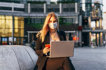 Businesswoman wearing mask using laptop and smart phone while sitting against building - MEUF01111
