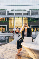 Geschäftsfrau mit Maske, die einen Laptop benutzt, während sie auf einem Sitz vor einem Gebäude sitzt - MEUF01112