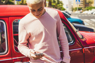 Smiling teenage boy standing in front of vintage car looking at smartphone - SIPF02170