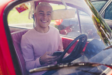 Portrait of smiling teenage boy sitting in vintage car with smartphone looking out of window - SIPF02171