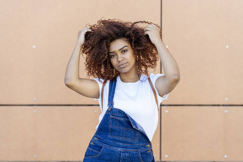 Confident young woman with hands in hair standing against wall - JSMF01562