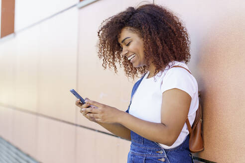 Glückliche junge Frau mit Afro-Haar, die ein Smartphone benutzt, während sie an einer Mauer in der Stadt steht - JSMF01565