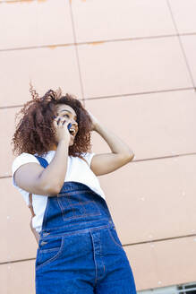 Fröhliche junge Frau mit Afro-Haar, die über ein Smartphone spricht, während sie an der Wand steht - JSMF01568
