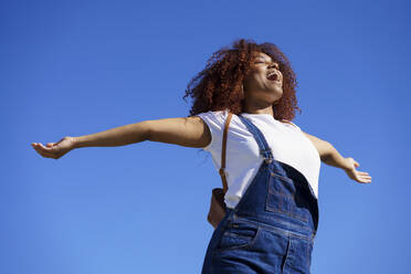 Sorglose Afro-Frau mit ausgestreckten Armen vor blauem Himmel - JSMF01570