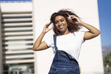 Lächelnde Afro-Frau, die Musik über Kopfhörer hört, während sie an einem Gebäude steht - JSMF01572