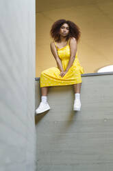 Junge afroamerikanische Frau in gelbem Kleid sitzt auf einer Stützmauer - JSMF01578