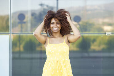 Happy young woman with hair in hair wearing yellow dress standing against wall - JSMF01580