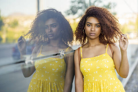 Serious afro woman wearing yellow dress standing by glass wall in city stock photo