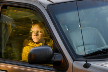 Boy sitting in car looking out passanger window tword setting sun - CAVF86590