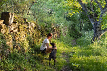 Frau streichelt und spielt mit ihrem Hund in einem Wald. - CAVF86655