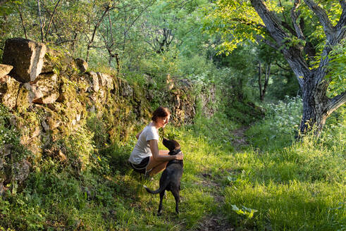 Frau streichelt und spielt mit ihrem Hund in einem Wald. - CAVF86655