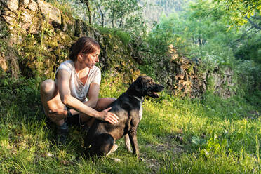 Frau streichelt und spielt mit ihrem Hund in einem Wald. - CAVF86664