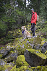 Männlicher Wanderer und flauschiger Hund stehen auf moosbewachsenen Felsen in den Bergen - CAVF86673