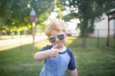 Young boy giving a thumbs up with sunglasses on standing in backyard - CAVF86688