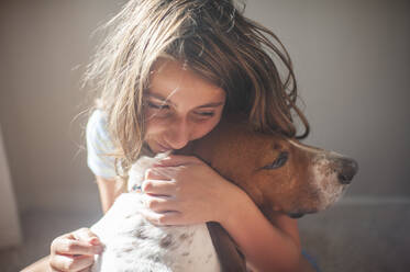 Happy Tween Girl hugging her Basset hound dog inside near a window - CAVF86689