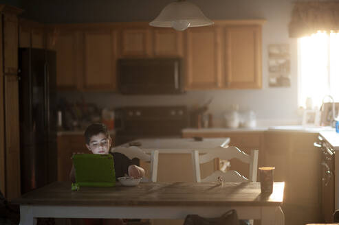 Kleiner Junge frühstückt, während er sein Tablet in der Küche beobachtet - CAVF86700
