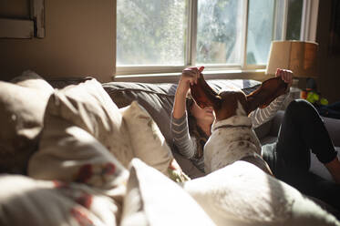 Mädchen sitzt auf der Couch mit Basset Hound Hund hält seine Ohren - CAVF86702