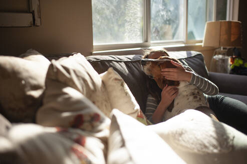 Girl hugging and kissing her dog on couch at home in pretty light - CAVF86704
