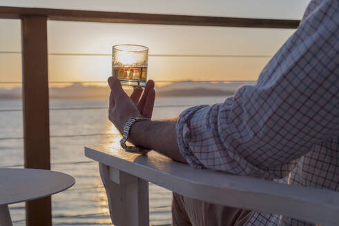 Mittelteil eines Mannes, der ein alkoholisches Getränk hält, während er bei Sonnenuntergang am Meer sitzt - EYF09073