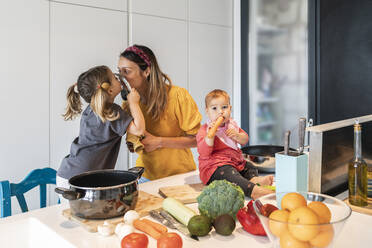 Mother and daughter kissing through lid while baby girl sitting on kitchen island at home - JAF00013