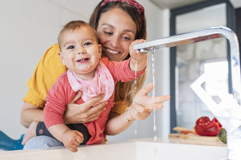 Smiling mother and cute baby girl playing with water falling from faucet in kitchen sink - JAF00014