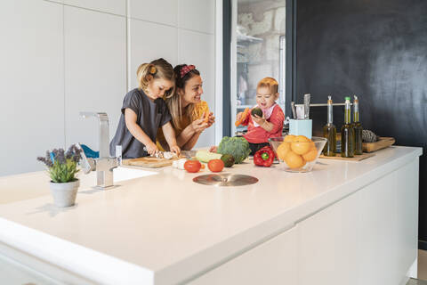 Ein kleines Mädchen sitzt auf einer Insel, während Mutter und Tochter in der Küche Essen zubereiten, lizenzfreies Stockfoto