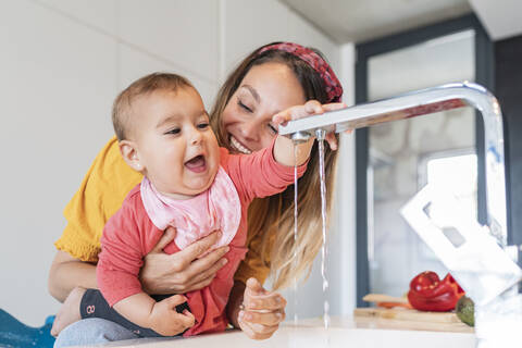 Nahaufnahme einer lächelnden Mutter, die ein süßes kleines Mädchen hält, das mit einem Wasserhahn in der Küche spielt, lizenzfreies Stockfoto