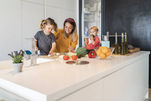Kleine Tochter sitzt auf einer Insel, während Mutter und Mädchen in der Küche Essen zubereiten - JAF00018