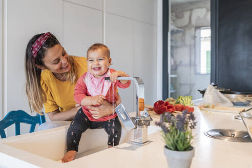 Smiling mother holding cute baby daughter playing with faucet in kitchen sink - JAF00019
