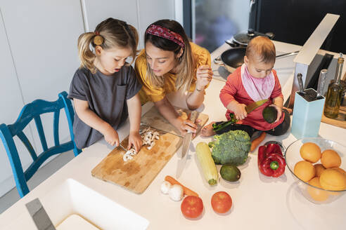 Baby girl sitting on island while mother looking at daughter chopping vegetables in kitchen - JAF00020