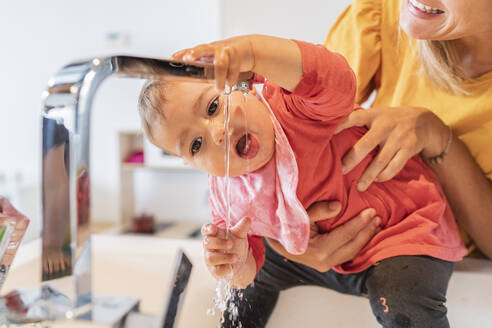 Nahaufnahme einer Mutter, die ein süßes kleines Mädchen hält, das Wasser aus dem Wasserhahn in der Küchenspüle trinkt - JAF00021