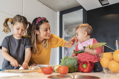Mutter spielt mit ihrer Tochter, während das Mädchen auf der Kücheninsel Gemüse schneidet, lizenzfreies Stockfoto