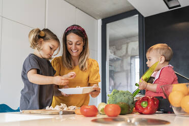 Mother and girl preparing food while baby daughter sitting on kitchen island - JAF00024