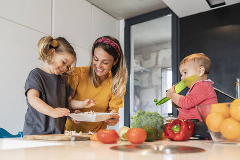 Smiling mother and girl preparing food while baby daughter sitting on kitchen island - JAF00025