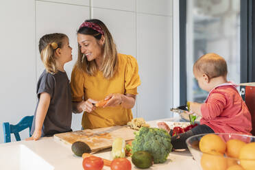 Lächelnde Mutter und Mädchen bereiten Essen zu, während die kleine Tochter auf der Kücheninsel spielt - JAF00026