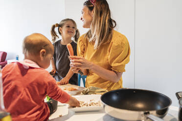 Lächelnde Mutter und Tochter schneiden Karotten, während das kleine Mädchen auf der Kücheninsel spielt - JAF00027