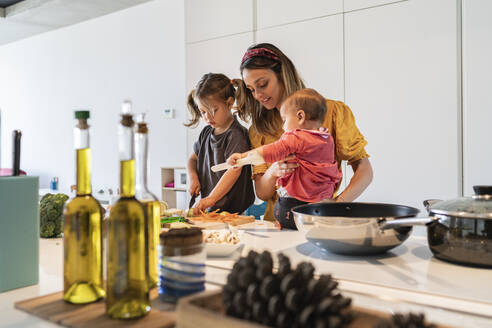 Mother holding baby girl while daughter cutting vegetables on kitchen island at home - JAF00028