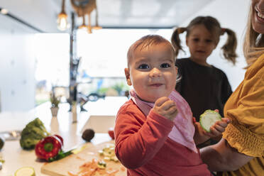 Nahaufnahme eines süßen kleinen Mädchens, das Zucchini isst, während Mutter und Tochter im Hintergrund arbeiten - JAF00029