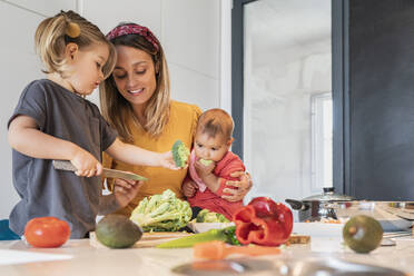 Mother holding baby girl while guiding daughter in cutting vegetables on kitchen island - JAF00031