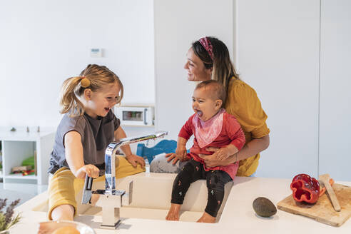 Mother and cheerful daughters playing with water in kitchen sink at home - JAF00032