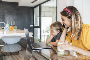 Mother with daughter looking at laptop while talking over smart phone in house - JAF00034