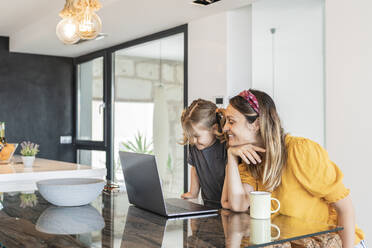 Smiling mother with daughter studying over laptop on table at home - JAF00035