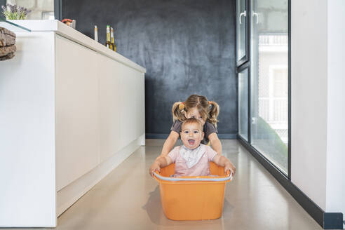 Girl pushing baby sister sitting in bucket on floor in modern kitchen - JAF00038