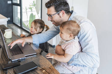 Vater arbeitet über Laptop auf Tisch mit Töchtern an seiner Seite zu Hause - JAF00039