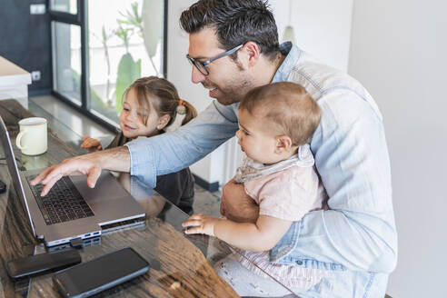 Father working over laptop on table with daughters by his side at home - JAF00039