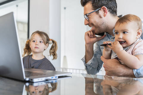 Man holding daughter talking over smart phone while girl looking at laptop on table stock photo
