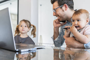 Mann hält Tochter im Gespräch über Smartphone, während Mädchen auf Laptop auf dem Tisch schaut - JAF00043