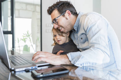 Father embracing daughter while working on laptop at home - JAF00044