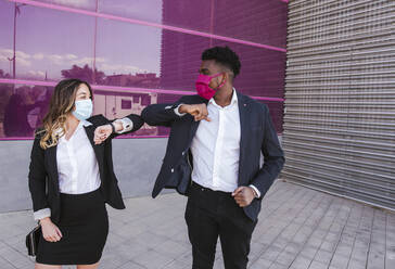 Business people wearing masks giving elbow bump while standing against modern building - LJF01629