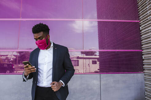 Businessman wearing mask using smart phone while standing against pink glass wall - LJF01635